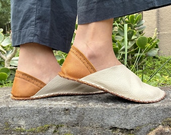 May include: A pair of slip-on shoes featuring a two-tone design with a cream-colored front section and a tan heel counter. The shoes are constructed with visible whipstitching along the sole and a decorative stitched pattern on the heel.
