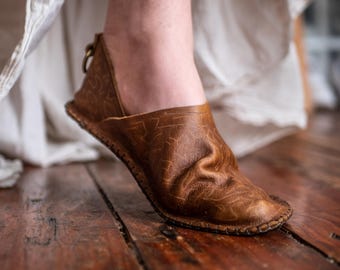 May include: A brown leather shoe with a textured surface and a stitched sole. The shoe has a rounded toe and a small loop on the back. The shoe is worn on a wooden floor.