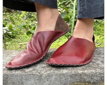 May include: A pair of deep red leather slip-on shoes featuring a soft, unstructured silhouette with a pointed toe and a dark brown heel counter. The shoes are constructed with visible black cross-stitching along the outer sole edge.