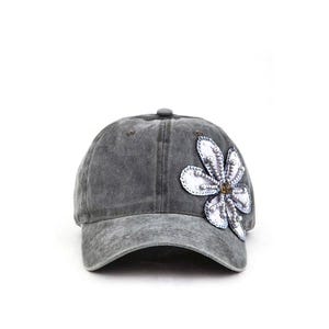 May include: A gray baseball cap with a distressed finish. The cap features a large, decorative flower on the side, embellished with white beads and a central brown gem. The cap has a curved brim and a button on top.