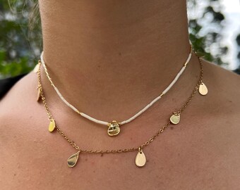 May include: Two necklaces worn around the neck. The top necklace is white with small gold beads. The bottom necklace is gold with teardrop-shaped pendants. The necklaces are gold in colour.