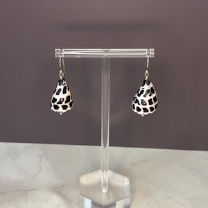 May include: A pair of drop earrings featuring natural cone shells with a distinct black and white spotted pattern. The shells are suspended from silver-toned metal hooks, showcasing a teardrop shape and a smooth, polished finish on the exterior.