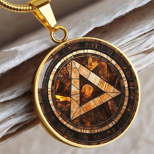 Puede incluir: Collar con colgante dorado con un diseño circular. El colgante presenta un patrón de mosaico en tonos marrones y beige, con un triángulo central y un pequeño círculo. La cadena del collar también es dorada.