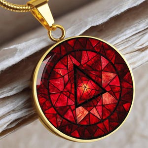 May include: A gold-colored pendant necklace with a circular, red mosaic design. The mosaic features a black triangle within a circle, set against a backdrop of red geometric shapes. The necklace has a gold chain.