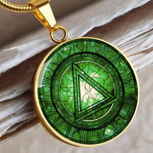 May include: Gold-toned pendant necklace featuring a circular, stained-glass-style design in shades of green. The pendant has a triangle within a circle, and is suspended from a gold chain. The necklace is displayed on a textured, off-white surface.