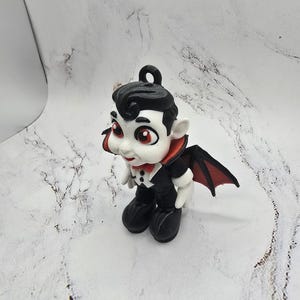 May include: A small, cartoon-style vampire figurine. The figure has a black hairstyle, a black suit, and a red bow tie. It has white skin, large red eyes, and small black wings. The figurine is designed to stand upright.