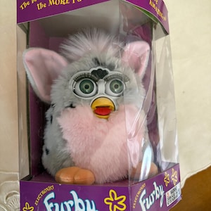 May include: A vintage Furby toy in its original packaging. The electronic toy has a gray and white body with pink accents, large green eyes, and a yellow beak. The packaging is purple with the Furby logo and text that reads, "The more you PLAY with me the MORE I do!"