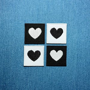 May include: Four square patches with heart designs. Two patches have a white heart on a black background, and two have a black heart on a white background. The patches are arranged in a square pattern against a blue denim background.