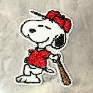 Op de afbeelding: Geborduurde patch met Snoopy in een honkbaluniform, met een knuppel. Snoopy draagt een rood shirt en pet, met een witte body en zwarte oren. De patch heeft een witte rand en staat op een lichtgrijze achtergrond.