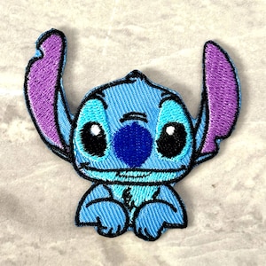 May include: Embroidered patch featuring the character Stitch from Lilo & Stitch. The patch is primarily light blue with dark blue and purple accents. Stitch has large black eyes and a friendly expression. The patch is designed for application on clothing or accessories.
