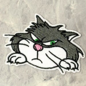 Puede incluir: Parche bordado con un gato de dibujos animados gruñón con pelaje gris, ojos verdes y nariz rosa. La cara del gato es blanca con contornos negros y tiene un borde blanco. El parche está diseñado para planchar o coser sobre tela.