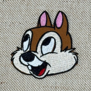 Könnte beinhalten: Gestickter Aufnäher mit dem Gesicht eines Cartoon-Chipmunks. Er hat einen braunen Kopf, eine beige Schnauze und rosa Ohren. Das Chipmunk hat große schwarze Augen, eine schwarze Nase und einen roten Mund. Auf beigem Stoffhintergrund.