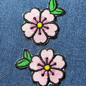 May include: Two embroidered patches featuring pink cherry blossoms with black outlines, yellow centers, and green leaves. The patches are set against a blue denim background, showcasing the detailed stitching and vibrant colors of the floral design.