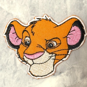 May include: Embroidered patch featuring the face of a cartoon lion cub. The patch has an orange body, pink ears with black outlines, and a white border. The lion cub has brown eyes and a smiling expression.