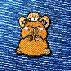 May include: Embroidered patch featuring a cartoon bear wearing a cowboy hat. The bear is brown with pink cheeks and black outlines. The patch is on a blue denim background.