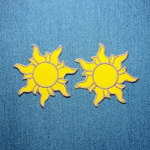 Può includere: Due toppe a forma di sole giallo con contorno viola e bordo dorato su sfondo denim blu. I soli hanno un centro circolare e raggi stilizzati.