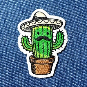 Puede incluir: Parche bordado con un cactus verde con sombrero y bigote negro. El cactus está en una maceta marrón. El parche tiene un borde blanco, sobre un fondo de mezclilla azul.