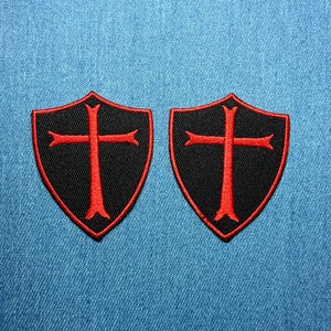 Może przedstawiać: Dwie czarne naszywki w kształcie tarczy z czerwonym krzyżem. Naszywki mają czerwoną obwódkę i są umieszczone na niebieskim tle z dżinsu. Idealne do odzieży lub akcesoriów.