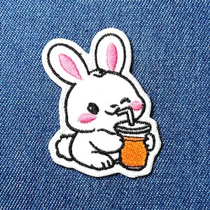 Op de afbeelding: Geborduurde patch met een witte cartoonkonijn met roze oren en wangen, drinkend uit een oranje beker met een rietje. De patch heeft een zwarte omtrek en een witte rand, tegen een blauwe denim achtergrond.