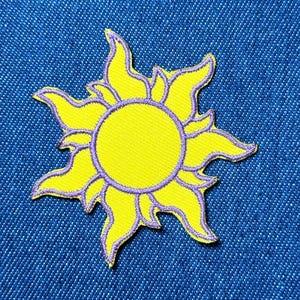 Puede incluir: Parche bordado con un diseño de sol amarillo con contornos y detalles morados. El sol tiene un centro circular y rayos estilizados. El parche está sobre un fondo de mezclilla azul, creando un contraste de colores.