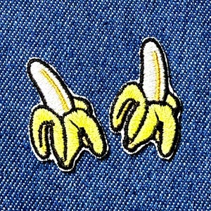 Peut inclure: Deux écussons brodés en forme de banane. Chaque écusson représente une banane jaune avec une peau blanche et un contour noir. Les écussons sont posés sur un fond en denim bleu.