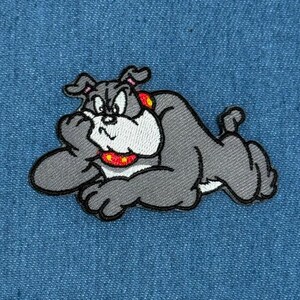 May include: An embroidered patch featuring a cartoon bulldog with gray fur, a white chest, and a red collar with yellow dots. The dog is depicted in a reclining position, resting its head on one paw with a grumpy facial expression and black outlines.