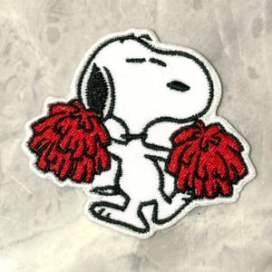 May include: Embroidered patch featuring a white cartoon dog with black ears and a black nose, holding two red pom-poms. The patch has a white border with black stitching, perfect for decorating clothing or accessories.