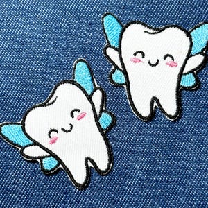 Könnte beinhalten: Zwei aufgestickte Aufnäher mit lächelnden Zeichentrickzähnen mit blauen Feenflügeln. Die Zähne sind weiß mit schwarzen Umrissen und rosa Wangen, auf blauem Denim-Hintergrund.