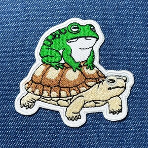 Op de afbeelding: Geborduurde patch met een groene kikker die op een bruine en beige schildpad rijdt. De patch heeft een witte rand en is geplaatst tegen een blauwe denim achtergrond. De kikker en de schildpad zijn gedetailleerd.