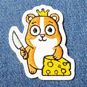 Peut inclure: Écusson brodé représentant un hamster de dessin animé portant une couronne et tenant un couteau, à côté d'un bloc de fromage jaune. Le hamster a une fourrure orange et blanche avec de grands yeux. L'écusson a une bordure blanche.