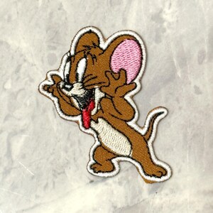 May include: Embroidered patch featuring Jerry Mouse from Tom and Jerry. The patch shows Jerry with a surprised expression, brown fur, pink ears, and a red tongue. The patch has a white border.