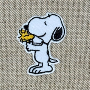 以下が含まれることがあります： ピーナッツのスヌーピーの刺繍パッチ。白地に黒のアクセント、黄色い鳥を抱えています。パッチは白い縁取りで、ベージュ色の生地の背景にあります。魅力的で懐かしいアイテムです。