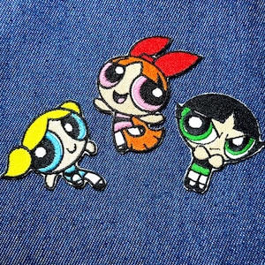 May include: Embroidered patches featuring the Powerpuff Girls characters Blossom, Bubbles, and Buttercup. Blossom has a red bow, Bubbles has blonde pigtails, and Buttercup has a black bob. The patches are on a blue denim background.
