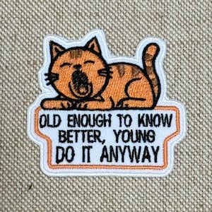May include: Embroidered patch featuring an orange tabby cat yawning, with the text "OLD ENOUGH TO KNOW BETTER, YOUNG DO IT ANYWAY" in black and orange. The patch has a white border and is on a tan fabric background.