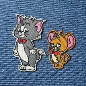 May include: Embroidered patches featuring cartoon characters Tom and Jerry. Tom is gray with pink ears and a red bow tie. Jerry is tan with pink ears and a red bow tie. Both patches are on a blue denim background.