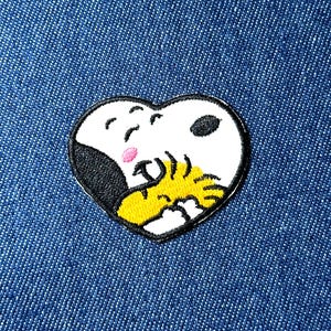 May include: Embroidered heart-shaped patch featuring Snoopy from Peanuts. The patch has a white background with black, yellow, and pink details. The patch is on a blue denim background.