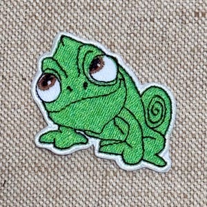 May include: Embroidered patch featuring a green chameleon with large brown eyes and a white outline. The patch has a detailed, textured appearance, and the chameleon is in a friendly pose. The patch is likely for clothing or accessories.