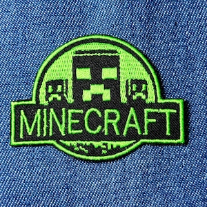Puede incluir: Un parche de Minecraft con un diseño negro y verde. El parche presenta el logotipo de Minecraft con la palabra "Minecraft" en letras verdes. El parche está sobre un fondo de mezclilla azul.