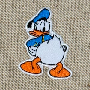 May include: Embroidered patch of Donald Duck in a walking pose. The patch has a white border, with Donald wearing a blue sailor shirt and hat, orange beak and feet, and a white backside. The patch is on a beige fabric background.