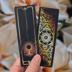 May include: Two black tarot cards with gold accents. One card features a celestial design with a dark circle and a cityscape. The other card has ornate gold patterns and the word "HYPERSPACE" and "RITUAL".