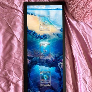 May include: Black framed artwork featuring three Pokémon trading cards set against a vibrant ocean backdrop. The artwork includes the text "cards are not included" at the top. The cards are displayed in a vertical arrangement, showcasing a beach and underwater scene.