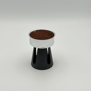 May include: A silver metal portafilter basket filled with dark brown coffee grounds sits atop a black espresso machine stand. The stand has a conical shape with vertical slots. The background is plain white.