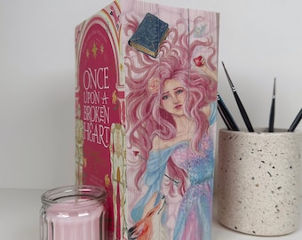 May include: A decorative wooden book with the title "Once Upon a Broken Heart" and a painting of a woman with pink hair and a fox. A small glass jar with a pink candle and a speckled ceramic container with paint brushes are also present.