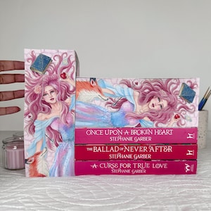 May include: A stack of three books with pink covers and titles: "ONCE UPON A BROKEN HEART", "THE BALLAD OF NEVER AFTER", and "A CURSE FOR TRUE LOVE" by Stephanie Garber. The books are next to a decorative bookend with a fantasy illustration of a woman, a fox, and a book.