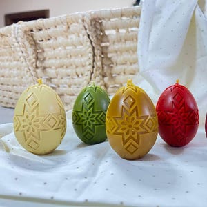 Puede incluir: Cinco huevos de Pascua decorativos hechos de cera de abejas, cada uno con un color diferente y un diseño de copo de nieve. Los huevos están dispuestos en una fila sobre una superficie blanca.