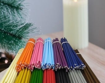 May include: Assortment of 14 colours of thin, long taper candles. The candles are bundled together and arranged in a rainbow pattern. A white pillar candle is in the background.