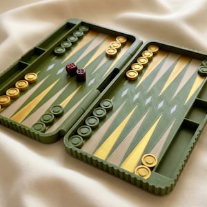 Könnte beinhalten: Geöffnetes Backgammon-Spielbrett in Olivgrün, Beige und Gold. Das Spielbrett zeigt die Spielfläche mit dreieckigen Punkten und Feldern für die Spielsteine. Zwei rote Würfel liegen auf dem Spielbrett, zusammen mit goldenen und grünen Spielsteinen.