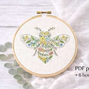 May include: A floral bee embroidery design in a wooden hoop. The bee is composed of colorful flowers and leaves. The text "PDF pattern + 6 hoop sizes" is visible on the bottom right.