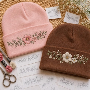 Puede incluir: Dos gorros bordados. Uno es rosa claro con flores rosas y hojas verdes, el otro es marrón con flores blancas y hojas verdes. Se ven hilos de bordar y tijeras.
