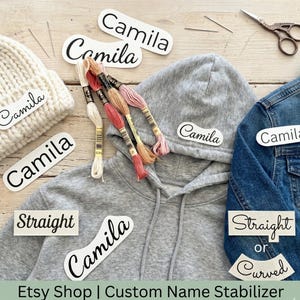 Op de afbeelding: Een assortiment gepersonaliseerde naamstabilisatoren met de naam "Camila" erop gedrukt. Items zijn onder meer een witte muts, een grijze hoodie en een spijkerjasje. Borduurgaren, naalden en een schaar zijn ook aanwezig.